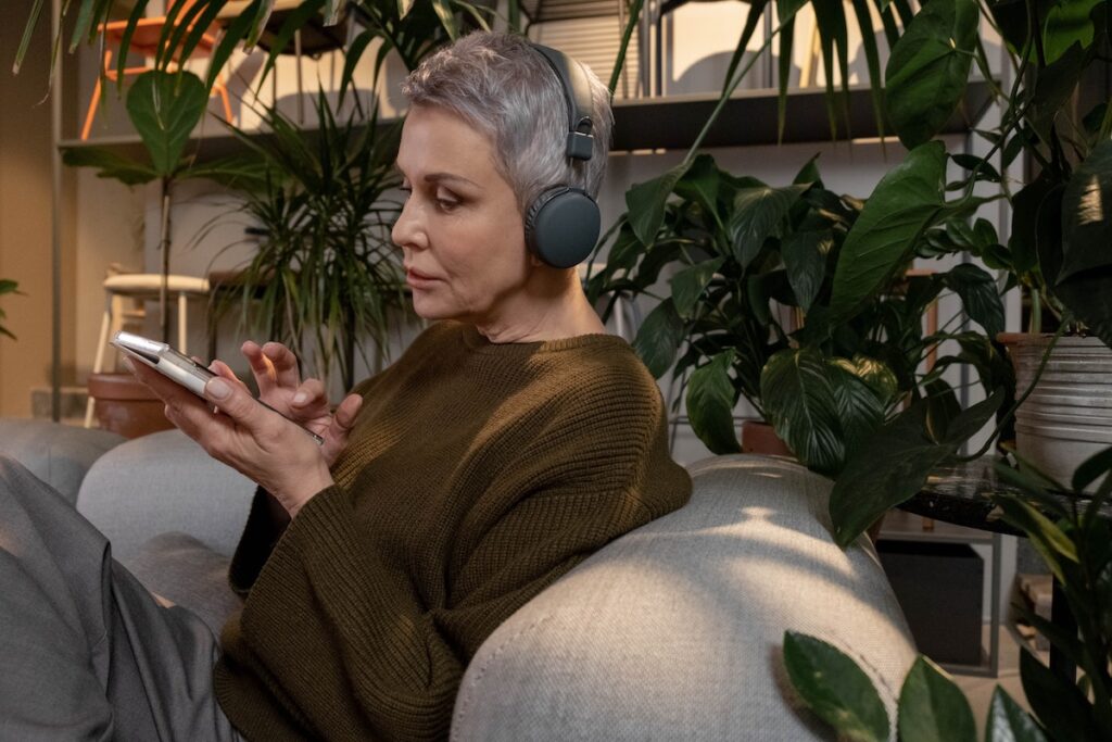 Older woman sitting on the couch listening to music with headphones on as one of her unique self-care ideas.