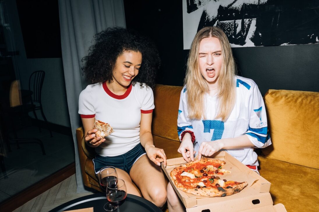 Two women eating pizza on a couch. 