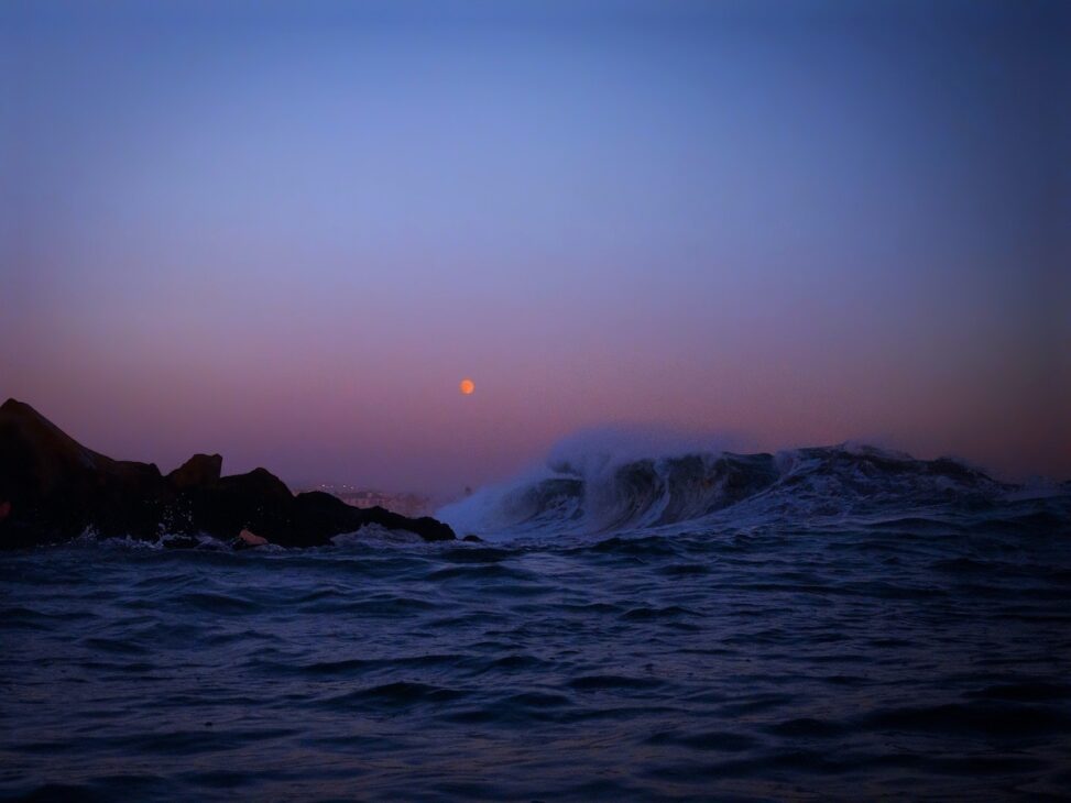 Waves crashing onto rocks with a moon in the sky. 
